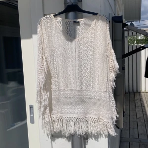 Vit spetsblus med fransar från Vila - Säljer en vit blus från Vila i spets med vackra virkade detaljer och fransar längs nederkanten och ärmarna. Blusen har lång ärm och rund halsringning, perfekt för en bohemisk look.
