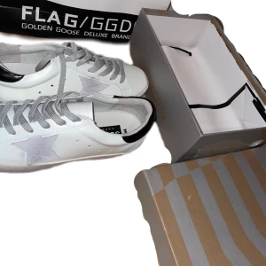 Golden Goose Superstar sneakers vita med grå detaljer - Golden Goose Superstar sneakers i vitt skinn med grå snörning och grå stjärndetalj på sidan. Klassisk låg modell med rund tå och platt sula. Märkeslogga på sidan och bak på hälen.