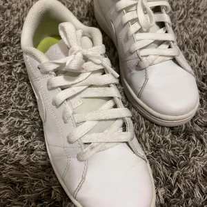 Vita sneakers med snörning - Säljer ett par klassiska vita sneakers från Adidas med snörning och rund tå. Skorna har en vit ovandel i syntetmaterial och vit sula. Innersulan är ljusgrön med svart Adidas-logga. Perfekta för en stilren vardagslook.