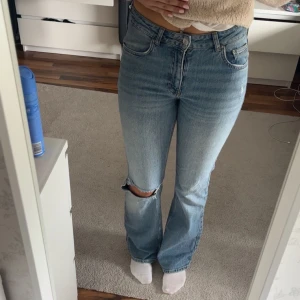 Ljusblå bootcut jeans med slitning - Säljer ett par ljusblå jeans med bootcut passform och hög midja. Jeansen har en tydlig slitning på ena knät och klassiska fem fickor. Perfekta för en avslappnad och trendig look. Är 172 cm