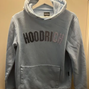 Ljusblå hoodie från Hoodrich - Säljer en ljusblå hoodie från Hoodrich med broderad logga i svart och vit på bröstet. Tröjan har huva, magficka och långa ärmar. Perfekt för en avslappnad stil.