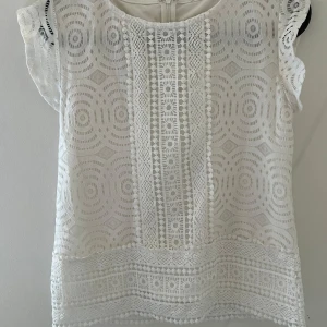 Vit spetsblus - Säljer en vit blus med vackert spetsmönster och korta ärmar. Blusen har dragkedja bak och fina detaljer längs framstycket. Perfekt nu till sommaren, vet ej vart den är ifrån💕