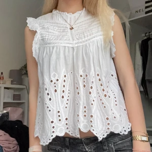 Vit blus med broderade detaljer och volang från Zara  - Säljer en söt vit blus från Zara med broderade hålmönster och volang vid axlarna. Blusen är ärmlös med spetsdetaljer och vågig nederkant. Perfekt för vår och sommar! 