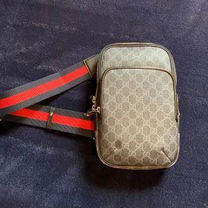 Gucci axelväska med monogram och röd-svart rem - Snygg axelväska från Gucci i grått med klassiskt GG-monogram. Väskan har en justerbar axelrem i svart och rött, dragkedja och ett ytterfack. 