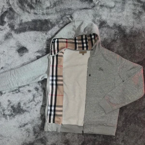 Grå zip hoodie från Burberry - Säljer en grå zip hoodie från Burberry. Mycket fint skick, inga defekter. Insidan av huvan har det ikoniska Burberry-rutiga fodret. Broderad logga på bröstet. Storlek L men passar som både S och M. Skriv ifall du har fler frågor!