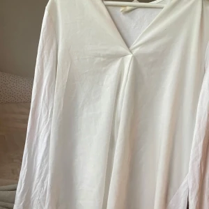 Vit långärmad v-ringad topp från Gina Tricot - Säljer en stilren vit långärmad topp från Gina Tricot med v-ringning och lätt omlott-detalj framtill. Perfekt att matcha med jeans eller kjol för en enkel och fräsch look.