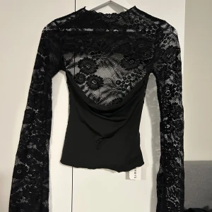 Svart spetsblus med lång ärm - Säljer en svart blus med lång ärm i transparent spets och blommigt mönster. Toppen har en snygg öppen rygg och figurnära passform. Perfekt för dig som vill sticka ut lite extra! Oanvänd