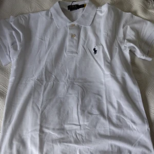 Vit pikétröja från Polo Ralph Lauren - Klassisk vit pikétröja från Polo Ralph Lauren med broderad logga på bröstet. Tröjan har korta ärmar, krage och knappar framtill. Tillverkad i bomull och har en slim fit passform.