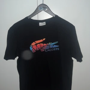 Svart t-shirt från Lacoste med färgglad logga - Säljer en svart t-shirt från Lacoste med en stor, färgglad krokodillogga på bröstet. T-shirten har rund halsringning och korta ärmar. Perfekt för dig som gillar klassiska märken med en twist.
