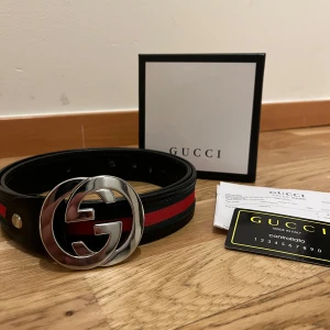 Gucci bälte - Säljer ett svart bälte från Gucci med bred röd och grön rand längs hela bältet. Stort, rundat spänne i silverfärgad metall med klassisk GG-logga. Box och kvitto medföljer och byten funkar också 