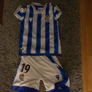 Real Sociedad fotbollströja och shorts - Säljer en blåvit randig fotbollströja från Real Sociedad med klubbmärke på bröstet och matchande vita shorts med blå detaljer och nummer 19. Klassisk V-ringning och korta ärmar. Perfekt för fotbollsälskare!