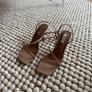 Beigea klackskor - Säljer ett par stilrena beige sandaler från NA-KD med smala remmar och fyrkantig tå. Skorna har en medelhög klack och öppen häl, perfekta för sommaren. Snygga till både jeans och klänning!