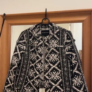 Svartvit mönstrad jacka från Who What Wear - Säljer en svartvit jacka från Who What Wear med grafiskt mönster och fransdetaljer vid axlarna. Jackan är croppad och har lång ärm, perfekt för att lyfta en outfit med sin unika design.