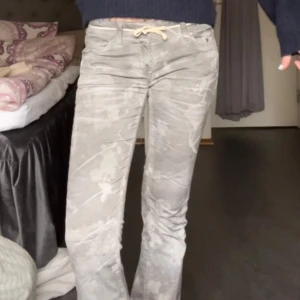 Grå bootcut jeans med batikmönster - Säljer ett par grå bootcut jeans med vitt batikmönster och snörning i midjan. Byxorna har en lågmidjad passform och är perfekta för dig som vill sticka ut lite extra. Vet ej vart dem är ifrån då jag köpte dem här på plick. 
