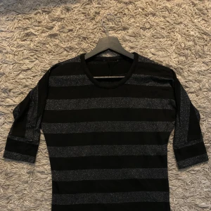 Svart och glittrig randig tröja - Säljer en svart tröja med breda horisontella glittriga ränder. Tröjan har rund halsringning och trekvartsärm. Perfekt för dig som vill ha något enkelt men ändå med lite extra glans.