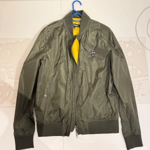 Olivgrön bomberjacka från J.Lindeberg - Snygg olivgrön bomberjacka från J.Lindeberg med gul insida. Jackan har dragkedja framtill, ribbade muddar och två fickor med tryckknappar. Perfekt för vår och höst, stilren och enkel att matcha.