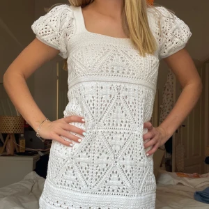 Vit virkad kortklänning med puffärm - Superfin vit virkad klänning med korta puffärmar och fyrkantig ringning. Klänningen har ett detaljerat hålmönster och sitter figurnära upptill med en lätt utställd kjol. Perfekt för sommarens alla tillfällen!