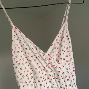 Vit klänning med körsbärsmönster - Supersöt vit kortklänning med tunna axelband. Klänningen har ett gulligt körsbärsmönster i rött och lätt rynkad midja för en flowy känsla. Perfekt för varma dagar och sommarhäng. Använt endast en gång.