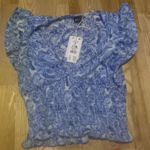 Blå smockad topp från Gina Tricot - Supersöt blå topp från Gina Tricot med paisleymönster och smockad midja. Toppen har volangdetaljer vid ärmslut och nederkant samt v-ringning. Perfekt för dig som gillar romantiska och trendiga plagg med extra detaljer. Aldrig använt den.