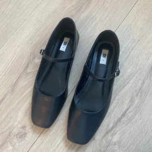 Svarta ballerinaskor - Svarta ballerinaskor från Gina Tricot med fyrkantig tå. Skorna har en tunn rem med spänne över foten och är gjorda i slätt syntetmaterial 