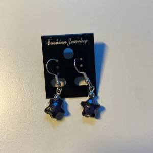 Handgjorda, Unika Silvriga örhängen med lila stjärnor *Endast 1 Kvar* - Ett par snygga örhängen med silvriga krokar och hängande lila stjärnformade berlocker. Perfekta för dig som vill ha något unikt och lekfullt i öronen. Stjärnorna har en glansig finish som fångar ljuset på ett coolt sätt.