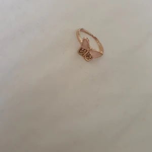 Guldfärgad ring med rosa fjäril - Superfin guldfärgad ring med en söt fjäril i ljusrosa på toppen. Fjärilen har glittriga detaljer på vingarna som ger extra bling. Perfekt accessoar för dig som gillar gulliga och trendiga smycken.