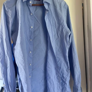 Ljusblå skjorta från Rose & Born - Säljer en klassisk ljusblå skjorta från Rose & Born. Skjortan har lång ärm, knappar framtill och är tillverkad i mjuk bomull. Perfekt för dig som gillar stilrena och tidlösa plagg. Snygg krage och enkel design som funkar till många outfits.