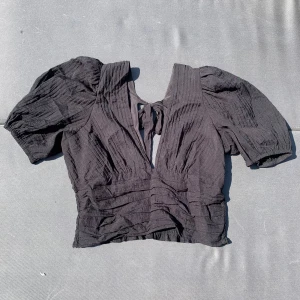 Svart blus med puffärm och knyt i ryggen - Säljer en svart blus med kort puffärm och snygg v-ringning. Toppen har knyt i ryggen och smockad midja för en skön passform. Materialet är lätt och har en krinklad struktur som ger cool textur. Perfekt för dig som gillar trendiga detaljer.