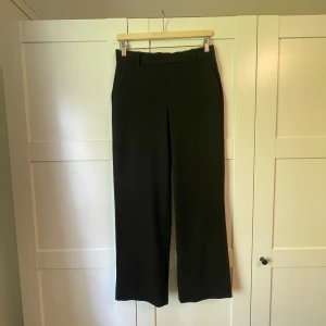 Svarta kostymbyxor från Vero Moda - Klassiska svarta kostymbyxor från Vero Moda i rak modell med hög midja och hällor för bälte. Byxorna har diskreta fickor i sidorna och elastisk midja bak för extra komfort. Perfekta för en clean och stilren look.