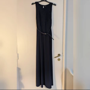 Mörkblå långklänning med tillhörande bälte/skärp - Oanvänd. Elegant och klassisk mörkblå långklänning med spetsliv och smalt bälte i midjan med gulddetalj. 
