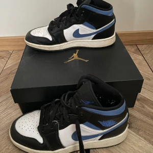 Nike Air Jordan 1 Mid svart/blå/vit - Säljer ett par Nike Air Jordan 1 Mid med svart, blå och vit färgkombination. Klassisk siluett med snörning, rund tå och vit sula. Ikoniska Jordan Wings-loggan på sidan och blå Swoosh. Äkta ifrån restocks och du får även med lådan på köpet. Unisex, storlek 36,5. Pris kan diskuteras! Köpta för 3000kr.