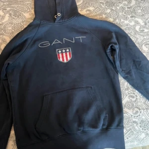 Mörkblå hoodie från GANT - Säljer en mörkblå hoodie från GANT med stor logga och sköld på bröstet. Tröjan har huva med snörning, magficka och långa ärmar. Perfekt för dig som gillar klassisk och sportig stil.