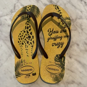 Gula Havaianas sandaler med giraffprint - Snygga flip-flops från Havaianas i gult med glittrig finish, bruna remmar och coolt giraffmönster samt palmblad. Texten 'You are giraffing me crazy' ger dem en lekfull vibe. Perfekta för sommaren och strandhäng.