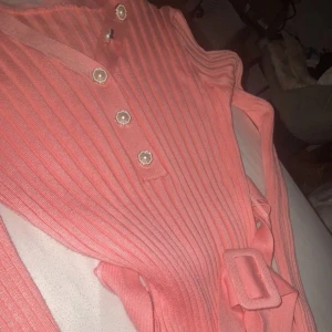 Ribbad rosa klänning med bälte - Supersöt ribbad rosa klänning med pärlknappar framtill och matchande bälte i midjan. Klänningen har en figurnära passform och är ärmlös, perfekt för en trendig look. Materialet känns mjukt och stretchigt, vilket gör den både bekväm och snygg.