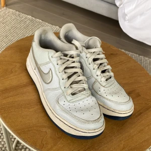 Nike Air Force 1 i ljusgrått skinn - Nike Air Force 1 sneakers i ljusgrått skinn med silverfärgade detaljer och blå sula. Klassisk rund tå, snörning och ikonisk swoosh på sidan. Perfekta för dig som gillar retrovibbar och streetstyle. Endast litet skavmärke på vänster sko. (Se bild 3)