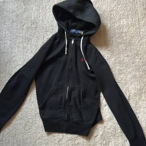 Svart hoodie från Polo Ralph Lauren XS - Svart hoodie från Polo Ralph Lauren i storlek XS. Klassisk design med dragkedja, huva med vita snören och röd broderad logga på bröstet. Mjuk bomullskänsla och kängurufickor framtill. Perfekt för en chill och stilren look.
