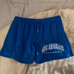Blå shorts från SHEIN Curve 1XL - Blå shorts från SHEIN Curve i storlek 1XL med vit text 'Los Angeles California' på ena benet. De har elastisk midja med snörning och är gjorda i mjuk bomull, perfekta för chill dagar eller träning. Klassisk loose fit och sportig vibe.