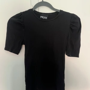 Svart och vit ribbad topp från Pieces - Två stilrena toppar från Pieces, en svart och en vit. Båda har ribbad struktur, puffiga korta ärmar och rund halsringning. Perfekt för dig som gillar enkel men trendig stil. Säljes tillsammans eller separat. Dem säljs i ett paket för 200kr.