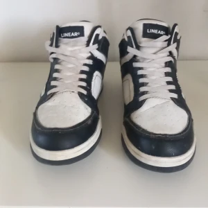 Svarta och vita sneakers från Linear - Snygga sneakers från Linear i svart och vitt med klassisk design. Skorna har snörning, rund tå och platt sula. Tillverkade i syntetmaterial med kontrasterande paneler och sportig vibe. Perfekta för dig som gillar streetstyle och enkel stil.