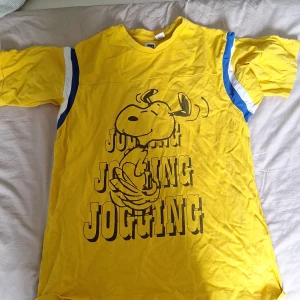 Gul Snoopy t-shirt Levi's x Peanuts S - Gul t-shirt från Levi's x Peanuts med stor Snoopy-tryck och texten 'JOGGING' på framsidan. T-shirten har korta ärmar med blå och vita ränder på axlarna. Tillverkad i mjuk bomull, perfekt för dig som gillar färg och coola prints.