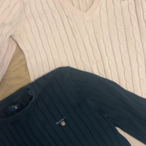 2 stickade GANT-tröjor, rosa & marinblå - Två snygga stickade tröjor från GANT i storlek S och mörkblåa i M (men sitter som S). En ljusrosa kabelstickad tröja med v-ringning och broderad logga på bröstet, samt en marinblå ribbstickad tröja med rund hals och broderad logga. Båda i mjuk bomull med klassisk passform. Prister gäller båda två!