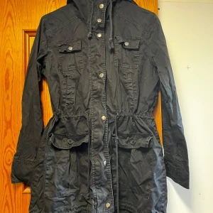 Svart parkajacka från Ellos - Svart parkajacka från Ellos med huva, dragsko i midjan och flera fickor framtill. Jackan har långa ärmar, knappar och dragkedja framtill samt snygga detaljer vid ärmslut och midja. Perfekt för dig som gillar en stilren och funktionell look. Jackan stryks och tvättas innan post☺️