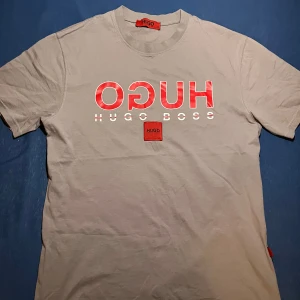 Hugo Boss t-shirt - Snygg grå t-shirt från Hugo Boss med stor röd och vit logga framtill. Klassisk rund halsringning och korta ärmar. Tillverkad i 100% bomull för en skön och luftig känsla. Perfekt för dig som gillar stilrena och trendiga plagg med tydlig branding.