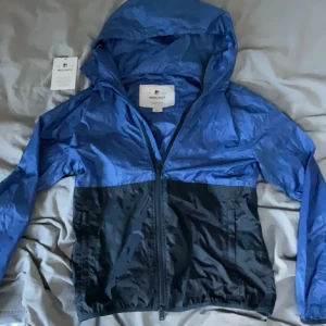 Blå och svart vindjacka från Woolrich - Snygg tvåfärgad vindjacka från Woolrich i blått och svart. Jackan har huva, hel dragkedja framtill och är tillverkad i lätt syntetmaterial. Perfekt för blåsiga dagar och enkel att packa ner i väskan. Klassisk streetstil med normal passform.