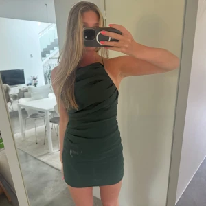 Mörkgrön kort klänning från Zara - Supersnygg mörkgrön kort klänning från Zara med smala axelband som korsas i ryggen. Klänningen har en tight passform och draperade detaljer framtill som ger en cool look. Perfekt för dig som vill sticka ut med en stilren och trendig outfit.