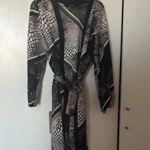 Långklänning med ormmönster och bälte - Snygg långklänning från Vero Moda med svart och beige ormmönster. Klänningen har långa ärmar, markerad midja med knytbälte och djup v-ringning. Perfekt för dig som vill sticka ut med ett coolt mönster och trendig look.