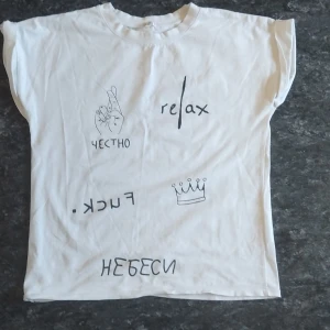 Vit t-shirt med tryck från Pudra - Vit t-shirt från Pudra med coola svarta tryck, bland annat texten 'relax', en krona, handillustration och rysk text. T-shirten har korta ärmar och en klassisk passform. Perfekt för dig som gillar unika prints och streetstyle.