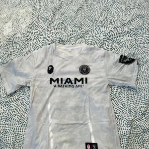 Inter Miami x BAPE fotbollströja vit - Snygg vit fotbollströja från Inter Miami i samarbete med A Bathing Ape. Tröjan har korta ärmar, svart tryck med MIAMI och BAPE-logga samt klubbmärke på bröstet. Lätt och bekvämt material, perfekt för dig som gillar streetwear och fotboll.