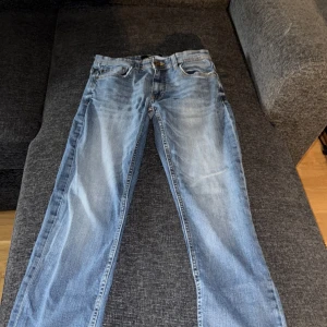 Ljusblå jeans från Smog, 30/32 - Säljer ett par ljusblå jeans från Smog i klassisk femficksmodell. Jeansen har raka ben, normal passform och snygga slitningar framtill för en avslappnad look. Tillverkade i bomull med kontrastsömmar och knappgylf. Det finns ett hål i fickan fram 