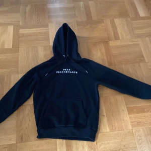 Svart hoodie från Peak Performance - Svart hoodie från Peak Performance med vit logga på bröstet. Klassisk modell med huva, snörning och stor magficka framtill. Mjuk insida och ribbade muddar vid ärmslut och nederkant. Perfekt för chill eller träning.